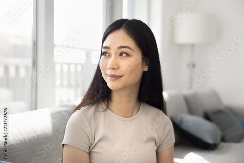 Happy dreamy beautiful Chinese girl sitting on sofa at home, looking at window away with dreamy thoughtful face, smiling. Positive young 20s Asian woman casual portrait