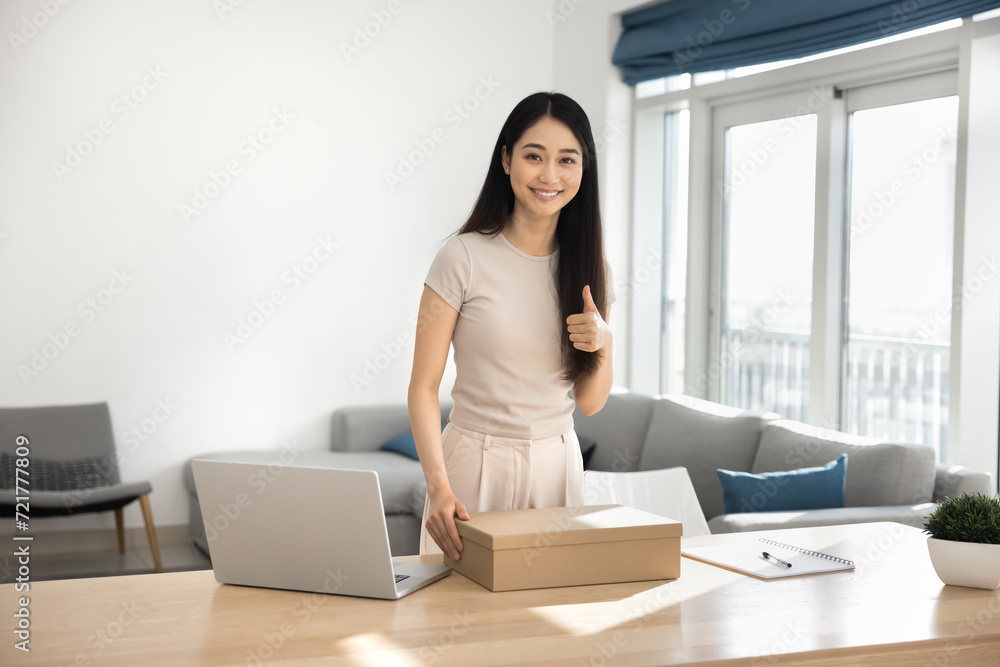 Happy young Asian customer girl receiving parcel from Internet store, holding cardboard box at home work table with laptop, looking at camera, showing like thumb up hand gesture, smiling Happy young Asian customer girl receiving parcel from Internet store, holding cardboard box at home work table with laptop, looking at camera, showing like thumb up hand gesture, smiling