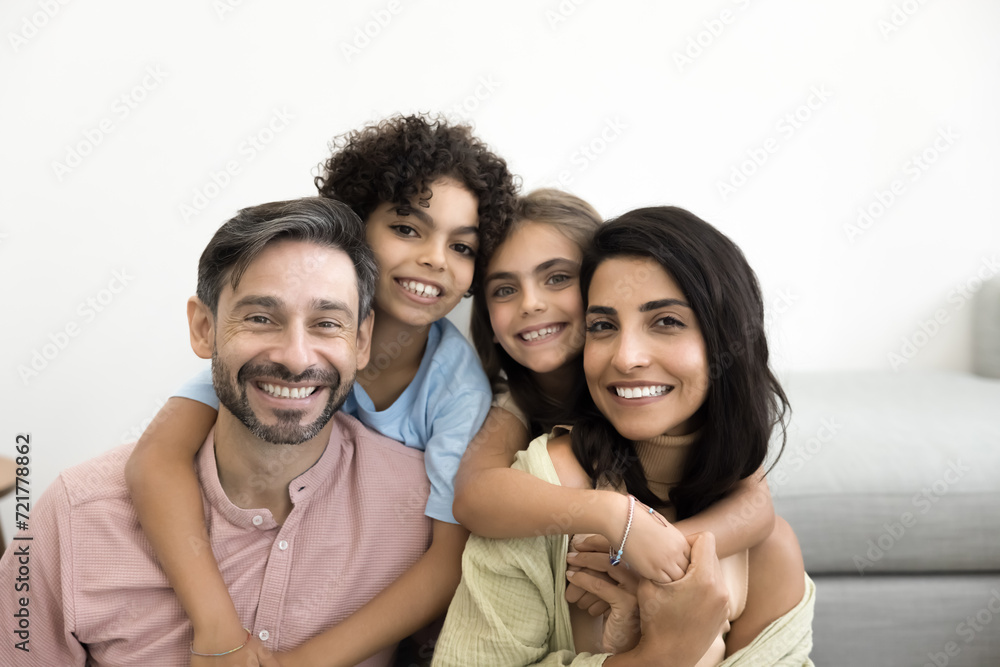 Cheerful Latin parents piggybacking preteen sibling kids, posing for family portrait at home, looking at camera with toothy smiles, laughing, enjoying childcare, parenthood