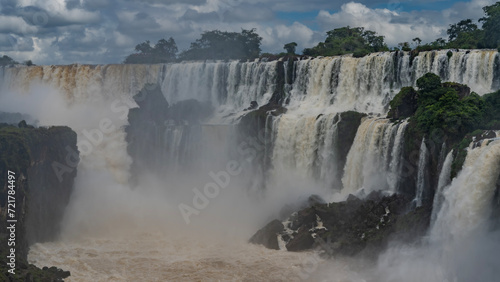 Famous tropical waterfalls. Streams, foaming, flow down the ledges of rocks into the riverbed. Splashes, fog. Lush green vegetation. Clouds in the blue sky. Iguazu Falls. Argentina.