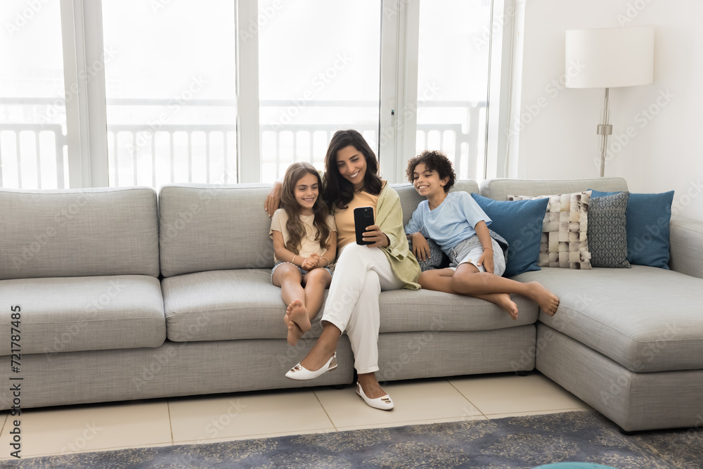 Cheerful young Latin mother and early teenage kids using internet technology, smart home application on smartphone, sitting on large comfortable couch, looking at screen, smiling, laughing