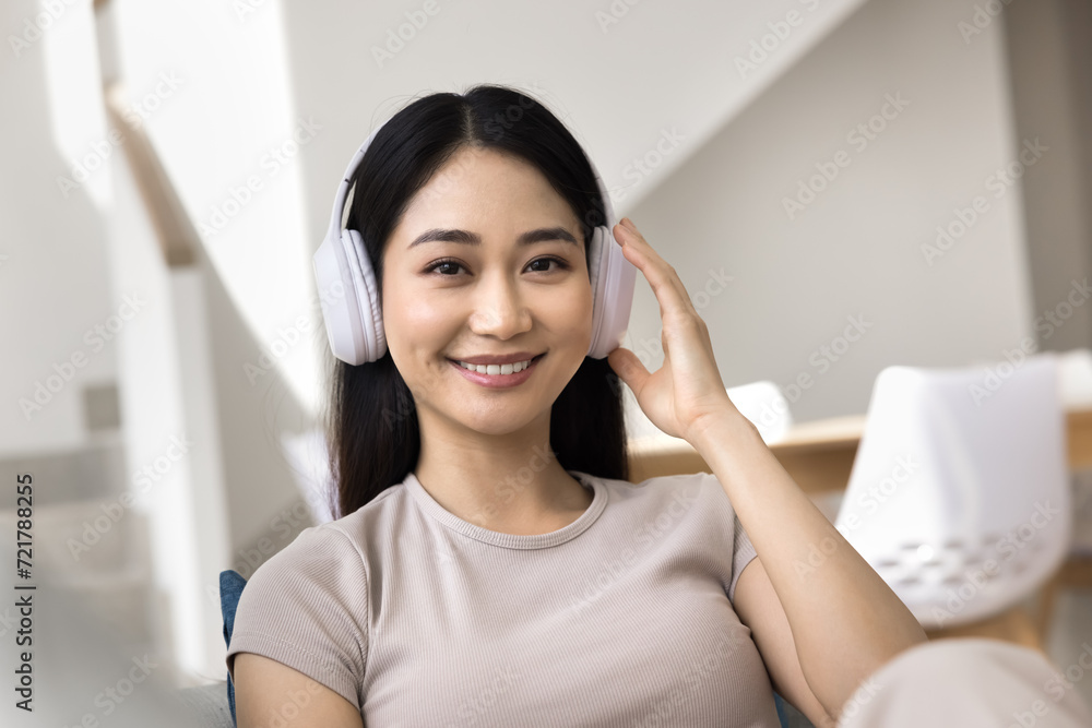 Happy Asian gadget user girl in headphones head shot portrait. Positive young woman touching big wireless earphones, looking at camera with toothy smile. Video call screen Happy Asian gadget user girl in headphones head shot portrait. Positive young woman touching big wireless earphones, looking at camera with toothy smile. Video call screen