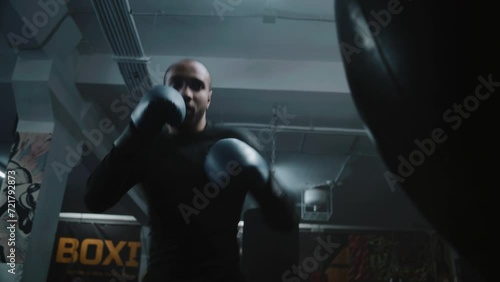 Confident African American fighter hits punching bag while training in dark boxing gym. Athletic man in boxing gloves exercises before fighting competition. Physical activity and intensive workout. photo