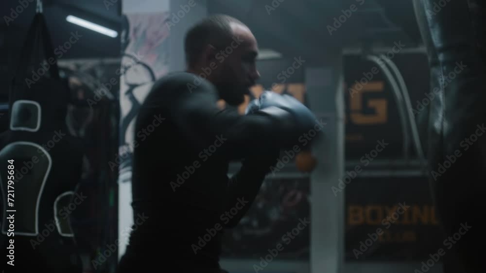 Focused African American fighter hits punching bag while training in dark boxing gym. Athletic adult man in boxing gloves exercises before fighting tournament. Physical activity and intensive workout. Focused African American fighter hits punching bag while training in dark boxing gym. Athletic adult man in boxing gloves exercises before fighting tournament. Physical activity and intensive workout.