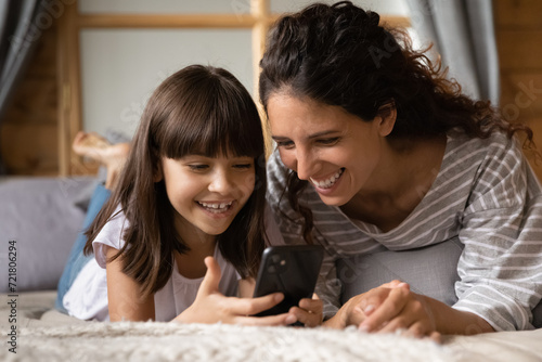 Pleasant leisure with cell. Excited young latin female mom or elder sister spend time lying on bed with cute little girl daughter using smartphone app. Mum and child enjoy pastime with phone together photo