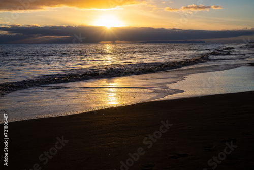 Fototapeta Naklejka Na Ścianę i Meble -  A sunset on Waimea Beach on Kauai reflects warmth onto gentle waves and a glow on the dark sand.