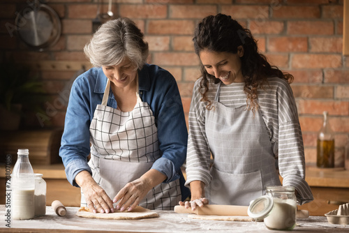 Doing cookies at home. Friendly older latin mother and daughter in law in kitchen aprons kneading rolling dough on table talk enjoy cooking together. Caring grownup child assist aged mom in baking pie photo