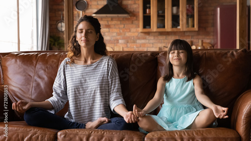 Mom and daughter yogis. Calm hispanic mother and little girl practice yoga exercise sit on sofa in lotus pose breath deep relax. Preteen child imitate elder sister nanny in doing meditative gymnastics