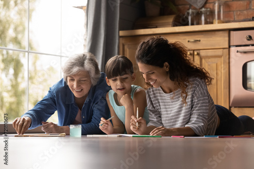 Family leisure. Friendly happy dynasty 3 latin females of different ages preteen girl kid older grandma young mom relax at home kitchen lying on warm floor drawing painting pictures with watercolors photo