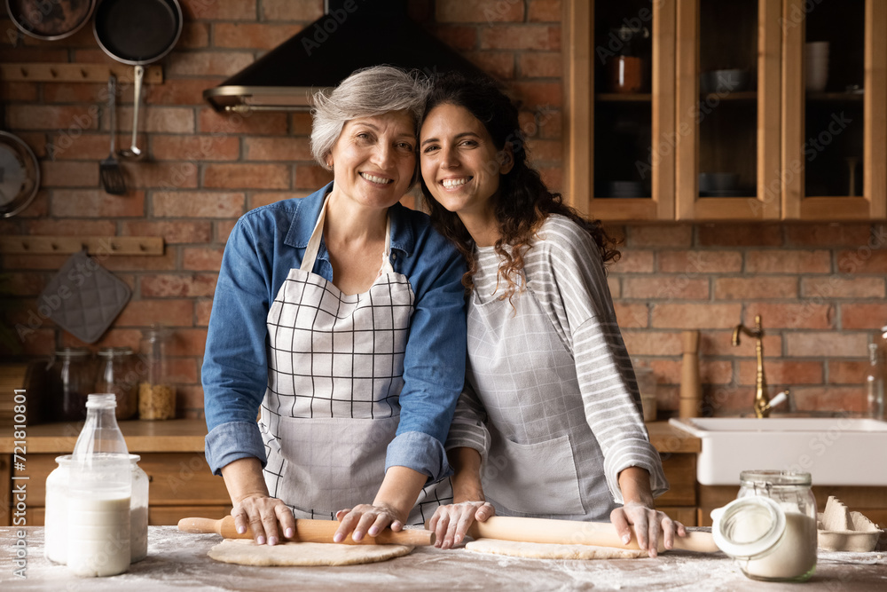 Fototapeta premium Portrait of two cheerful latina women retired mother young adult daughter wear aprons hug at kitchen table look at camera distracted of rolling dough. Senior granny grown grandkid cook homemade bakery