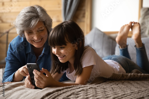 Happy preteen kid granddaughter rest on large bed with loving retired hispanic granny buying toys on ecommerce web site play cellphone game in online app. Mature babysitter use phone with little girl photo