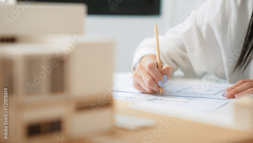 Closeup of beautiful young architect engineer hand drawing, drafting blueprint with blueprint and house model placed on working table at modern office. Focus on hand. Blurring background. Immaculate.