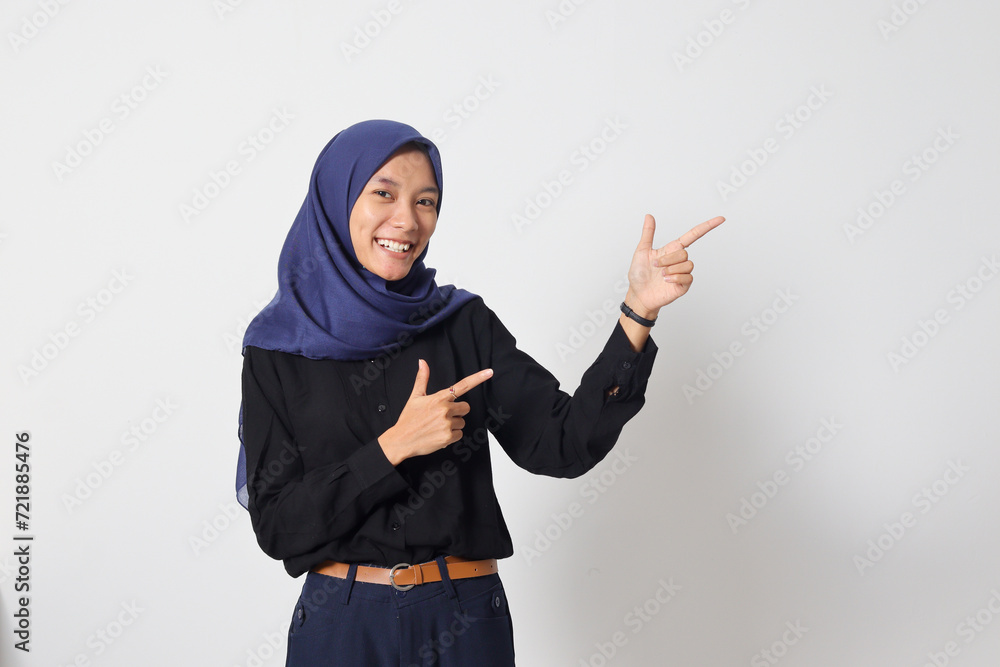 Portrait of excited Asian hijab woman in casual shirt promoting product, pointing finger to the side. Advertising concept. Isolated image on white background Portrait of excited Asian hijab woman in casual shirt promoting product, pointing finger to the side. Advertising concept. Isolated image on white background