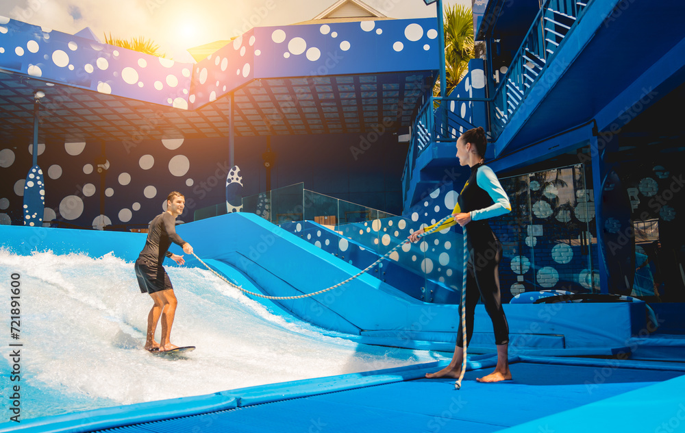 Young man surfing with trainer on a wave simulator at a water amusement park