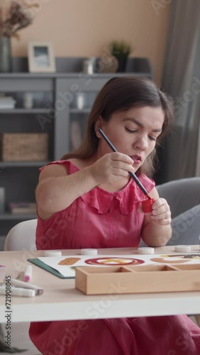 Vertical medium shot of young Caucasian woman with dwarfism wearing nice pink dress sitting at desk in modern living room at daytime and painting with colorful gouache on sheet of paperVertical medium photo