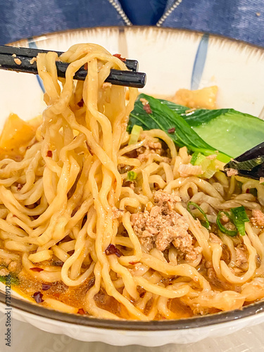 Eating chinese noodle soup topped with dumplings, chicken or beef meat, meatballs, vegetables served in ceramic bowls on a wooden table. photo