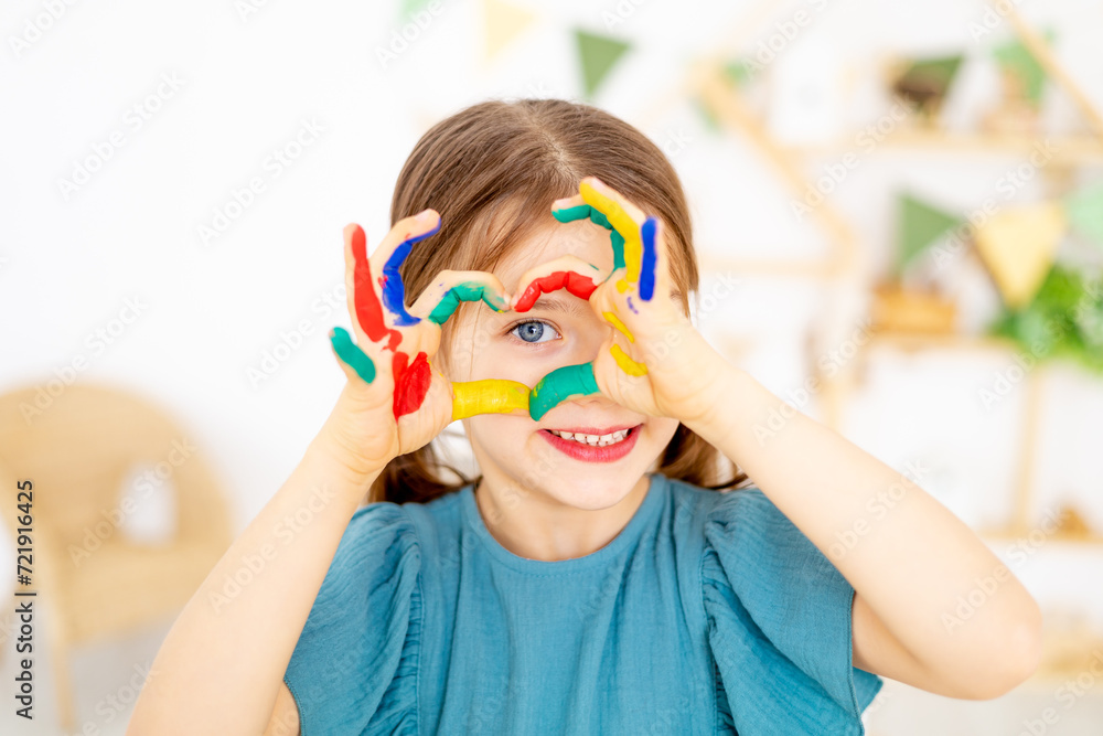 Naklejka premium cute little baby girl with colorful painted stained hands in finger paints shows a heart in the nursery and learns to draw or fooling around, art lessons with pencils and paints, close-up