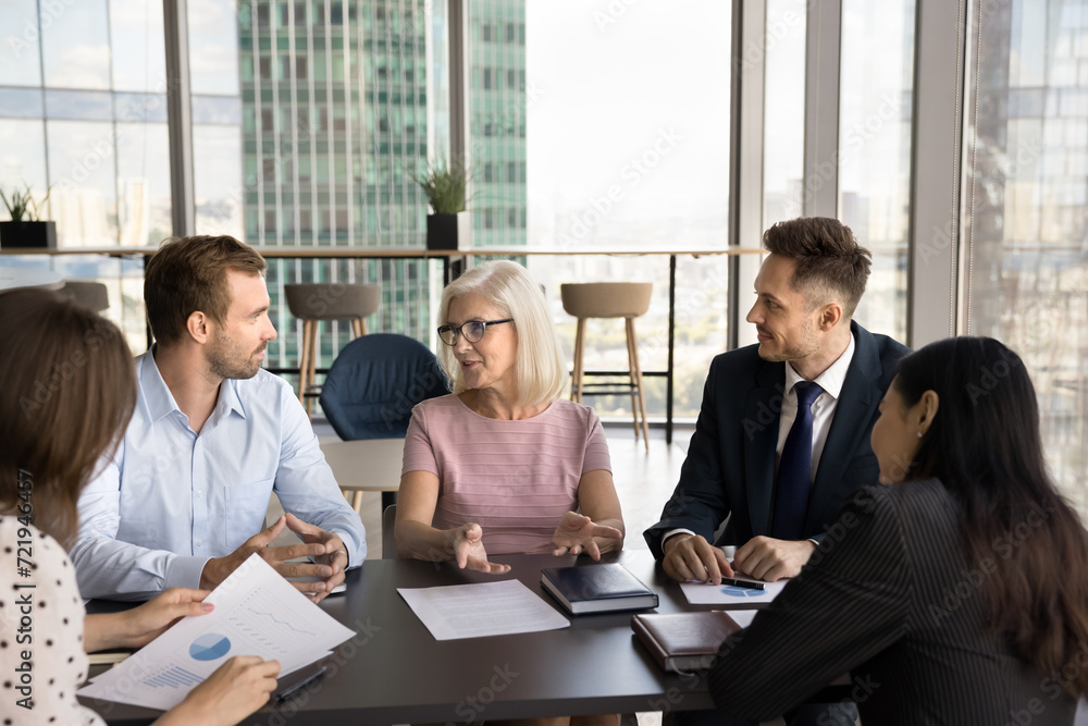 Positive senior business leader woman meeting with younger colleagues at negotiation table, offering ideas for brainstorming. Senior boss talking to listening coworkers, instructing team Positive senior business leader woman meeting with younger colleagues at negotiation table, offering ideas for brainstorming. Senior boss talking to listening coworkers, instructing team