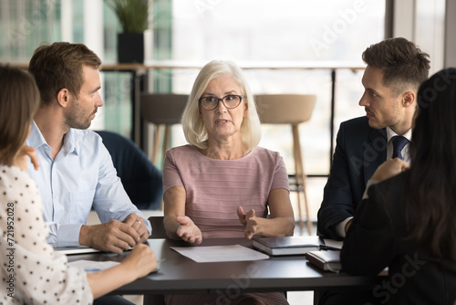 Confident serious elder female boss motivating team for teamwork, collaboration, talking to colleagues on corporate meeting, discussing project management, brainstorming with coworkers