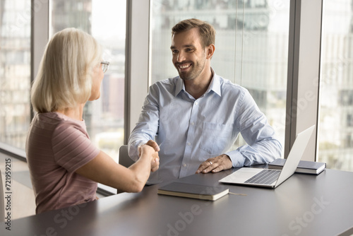 Happy young business professional man shaking hands with elder colleague woman, discussing collaboration at workplace table, giving handshake after talking on partnership, retting agreement photo