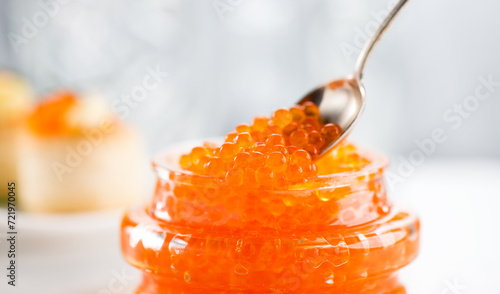Red Caviar in a spoon, fish roe in a glass jar. Close-up of salmon fish roe caviar on served table. Delicatessen. Texture of fresh trout caviar. Backdrop. Seafood. 