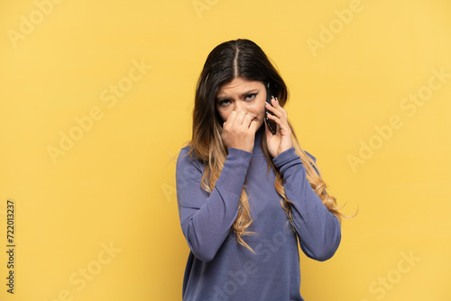 Young Russian girl using mobile phone isolated on yellow background having doubts