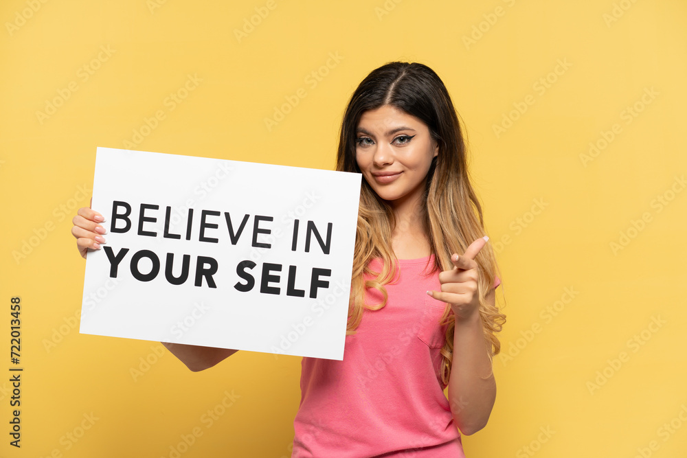 Young Russian girl isolated on yellow background holding a placard with text Believe In Your Self and pointing to the front Young Russian girl isolated on yellow background holding a placard with text Believe In Your Self and pointing to the front