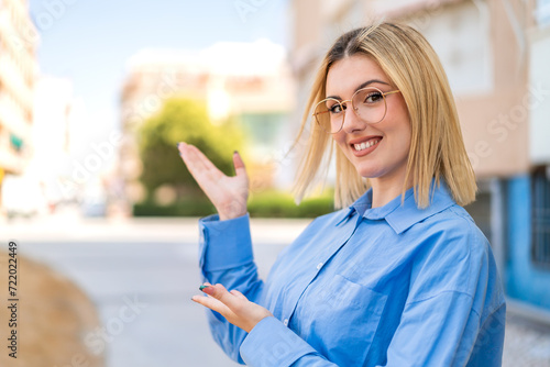 Young pretty blonde woman at outdoors With glasses and presenting something