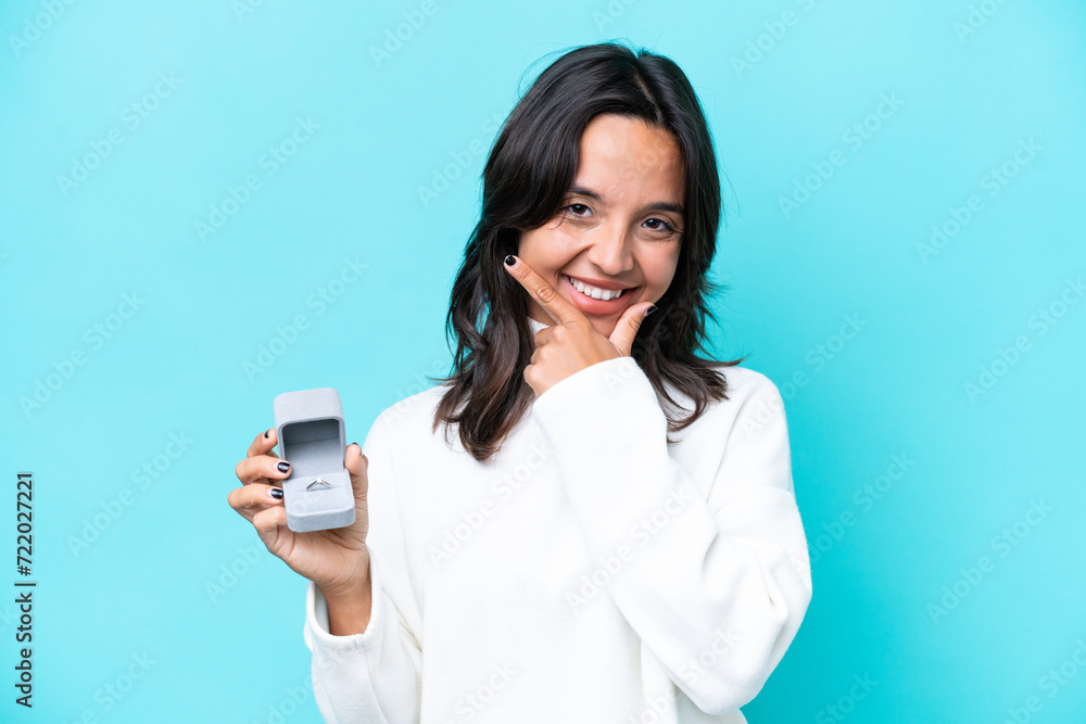 Young hispanic woman holding a engagement ring isolated on blue background happy and smiling Young hispanic woman holding a engagement ring isolated on blue background happy and smiling