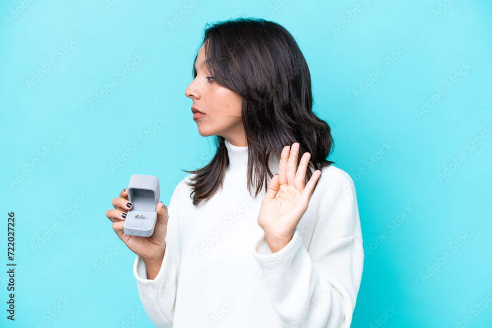 Young hispanic woman holding a engagement ring isolated on blue background making stop gesture and disappointed Young hispanic woman holding a engagement ring isolated on blue background making stop gesture and disappointed