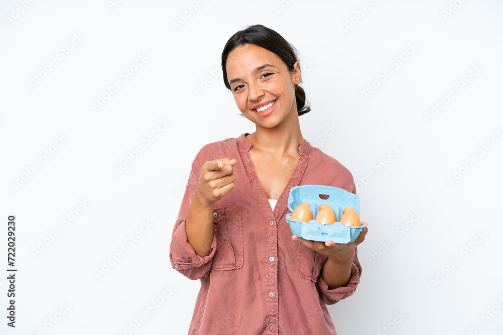 Young hispanic woman holding eggs isolated on white background pointing front with happy expression Young hispanic woman holding eggs isolated on white background pointing front with happy expression
