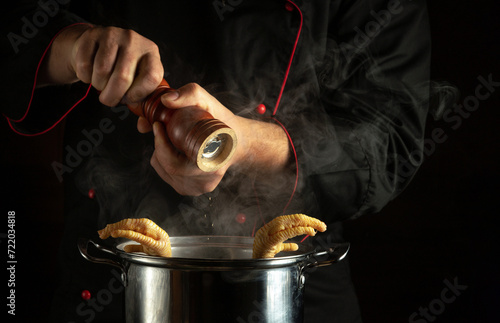 A cook in a black uniform adds pepper to a pot of boiling chicken legs. The concept of preparing a delicious and rich soup in the kitchen photo