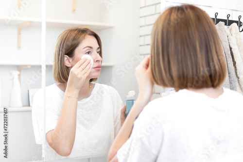 Attractive woman looking at mirror and applying moisturizing cream on cheeks in bathroom 