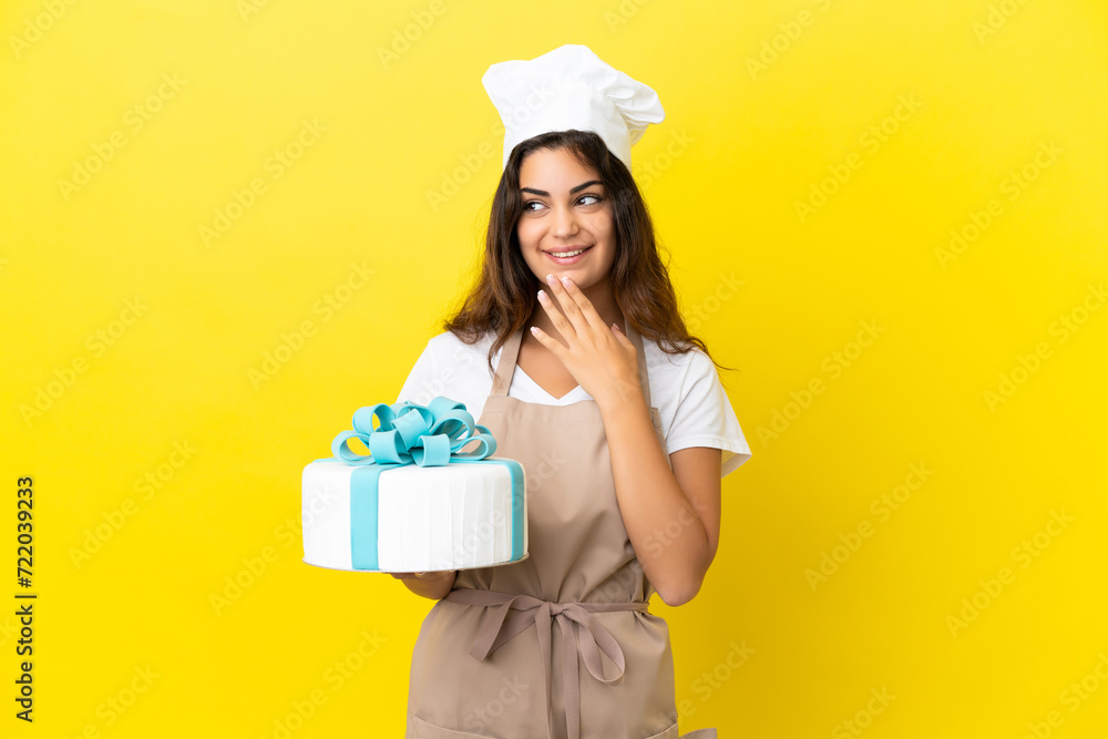 Young caucasian pastry chef woman with a big cake isolated on yellow background looking up while smiling Young caucasian pastry chef woman with a big cake isolated on yellow background looking up while smiling