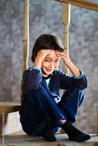 Fotografie Fake tears of little boy, 7 years old, of European appearance, sitting on step of wooden staircase in private two story house