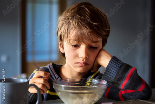 Dissatisfied Caucasian young boy is having breakfast while sitting in front of transparent bowl of porridge in dimly lit kitchen. photo