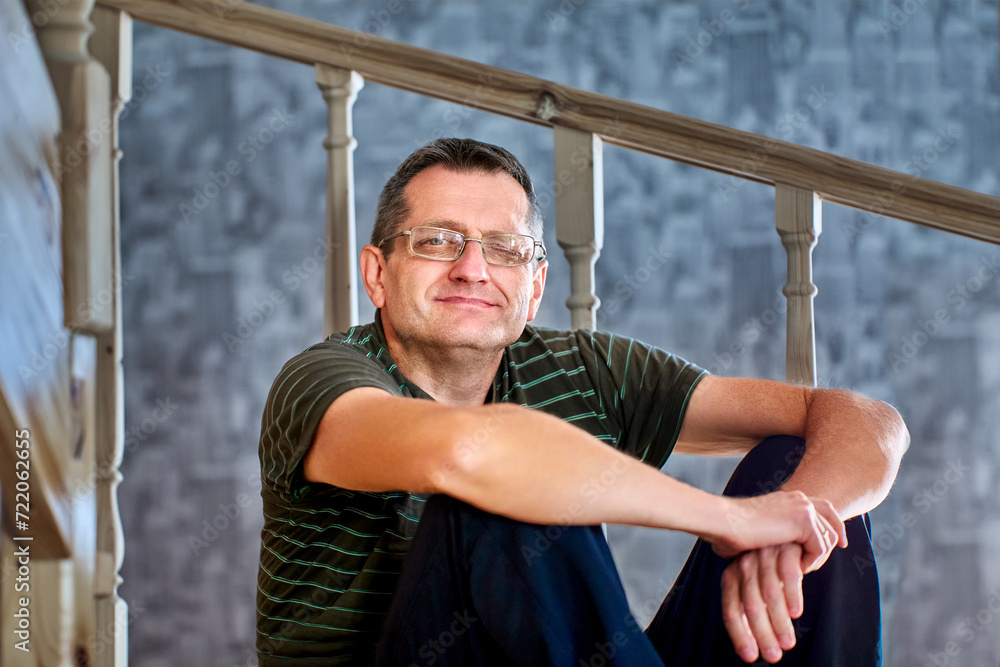 Obraz premium Ptosis of left eyelid in mature European man sitting on an interfloor wooden staircase at home.