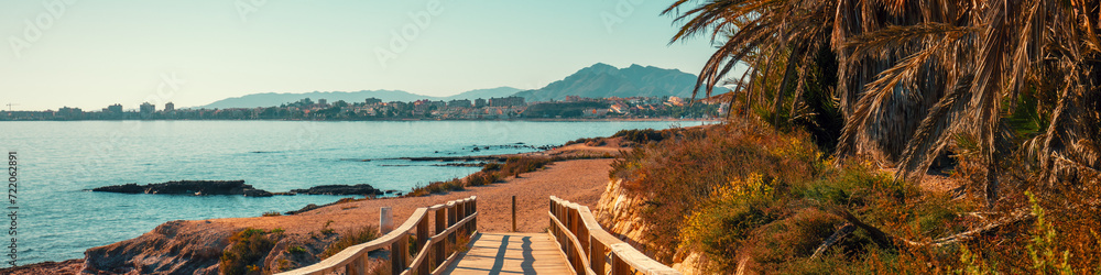 Seascape on a sunny day with a clear blue sky. Palm trees on the beach. Boardwalk to the beach. Mazarron, Murcia, Spain. Horizontal banner