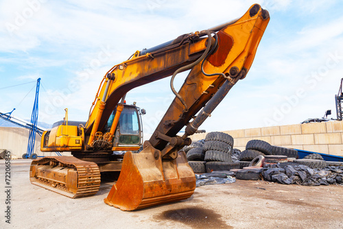 A crawler excavator stands on the port grounds next to a pile of old tires.