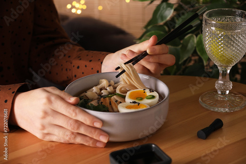 Woman eating delicious ramen with chopsticks at wooden table indoors, closeup. Noodle soup