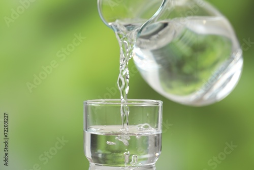 Pouring fresh water from jug into glass against blurred green background, closeup