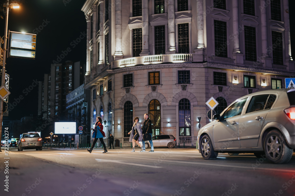 Fototapeta premium A vibrant nighttime cityscape with business partners walking at a crosswalk. The scene captures urban life, street illumination, and the energy of evening activities.