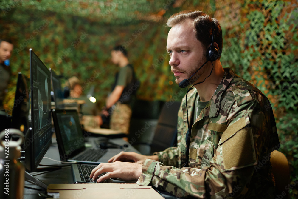 Young serious male operator with headset looking at graphic data on computer screen and keeping fingertips on keys of keyboard