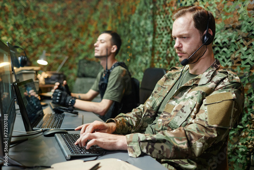 Serious young male operator in camouflage uniform typing on computer keyboard and looking at screen against colleague with walkie-talkie