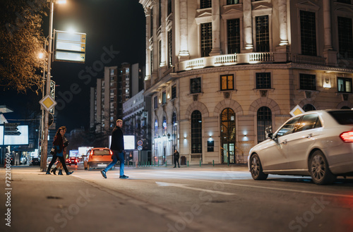 Two pedestrians casually crossing a well-lit city street at night with cars passing by and illuminated buildings in the backdrop. © qunica.com