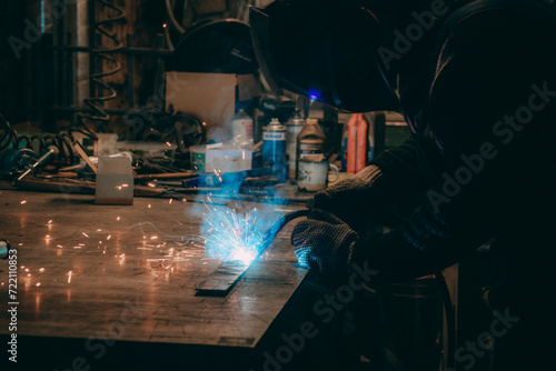 Master is doing welding at his workplace in the workshop, while sparks are flying all around, they are wearing a protective helmet.