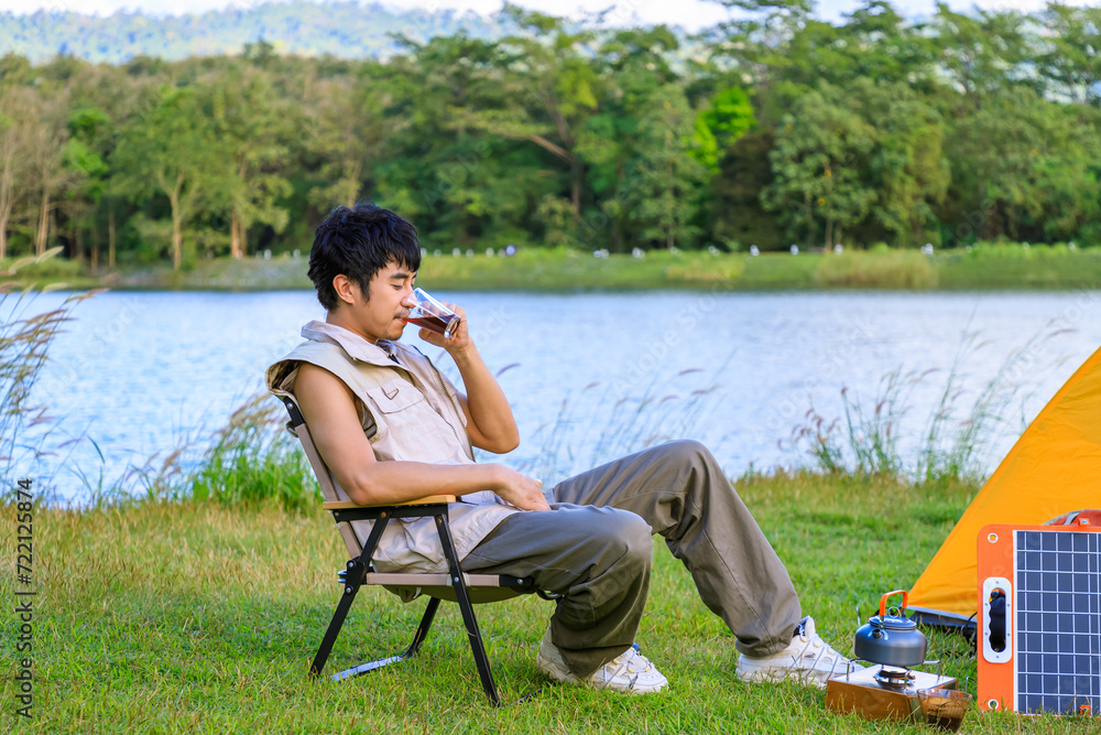 Asian male man sitting on chair, drinking coffee and feeling relax with camping tent beside lake and mountain, with mobile solar cell.