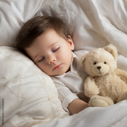 Happy baby boy lying in bed with closed eyes. Beautiful infant sleeping in bed wrapped in a white blanket. Cute toddler boy dreaming in bed with a big teddy bear.