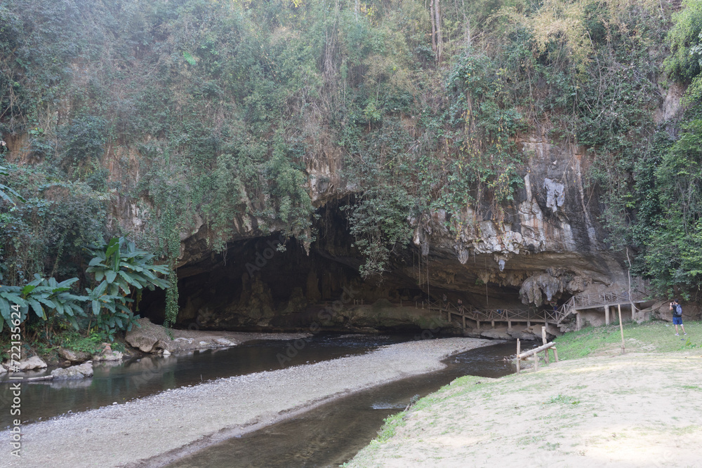 A serene natural landscape featuring a large cave entrance next to a gentle river with verdant foliage and a walking path. A serene natural landscape featuring a large cave entrance next to a gentle river with verdant foliage and a walking path.