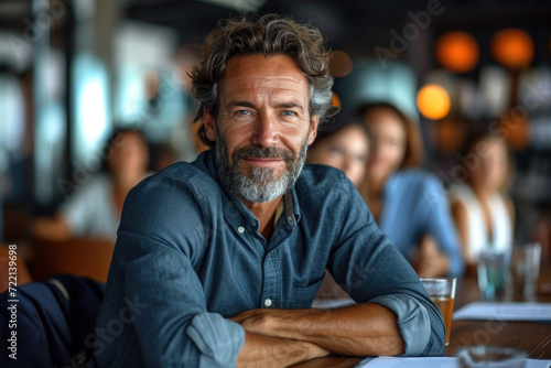 Successful business man sitting in a boardroom with his team in the background, looking at camera smiling, bokeh